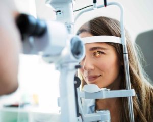 Während einer Augenuntersuchung stützt eine junge Patientin entspannt lächelnd ihr Kinn in das medizinische Gerät und schaut aufmerksam geradeaus. In ihrem Auge sind Lichtpunkte zu erkennen.