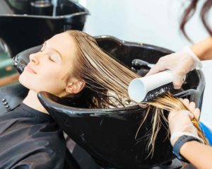 In einem Friseursalon lehnt eine junge Frau mit langen Haaren und geschlossenen Augen an einem Haarwaschbecken und bekommt die Haare gewaschen.