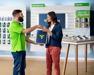 Im Freenet Store überreicht ein bärtiger Mitarbeiter im grünen T-Shirt einer Kundin eine blaue Einkaufstüte. Im Hintergrund sind Smartphones ausgestellt.