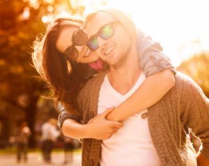 Im Sonnenschein umarmt eine junge Frau einen jungen Mann von hinten und schaut lachend über seine Schulter. Beide tragen modische Sonnenbrillen.