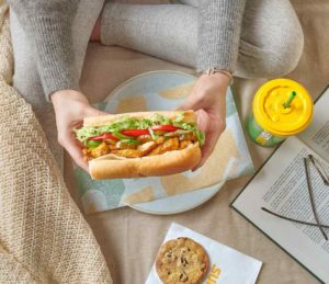 Eine Frau in legerer Kleidung sitzt auf dem Boden. Vor ihr stehen ein Teller und ein Getränkebecher von Subway. Sie hält ein üppig belegtes Sandwich in Händen. Vor ihr liegt zudem ein heller Cookie auf einer Serviette. Eine Frau in legerer Kleidung sitzt auf dem Boden. Vor ihr stehen ein Teller und ein Getränkebecher von Subway. Sie hält ein üppig belegtes Sandwich in Händen. Vor ihr liegt zudem ein heller Cookie auf einer Serviette.