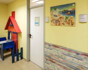 In der Logoädie-Praxis hängt ein buntes Bild mit Bärchen an einer gelben Wand. In einer Kinderecke steht ein Tisch mit blauen Stühlen. An der Wand ist eine bunte Hausfront in Kindergröße als Dekoration montiert.