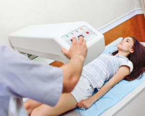 Eine junge Frau in Shorts und T-Shirt liegt auf einem Behandlungstisch. Eine Mitarbeiterin in grauem Kittel aktiviert ein Gerät, welches sich über der Hüfte der Patientin befindet.