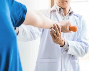Eine Person in blauem T-Shirt macht Armübungen mit einem kleinen Gewicht. Ein Arzt im weißen Kittel untersucht den ausgetreckten Arm.