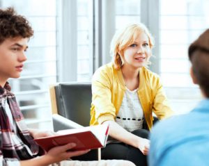 Vor einer Fensterfront sitzt eine junge Frau in gelber Jacke vorgebeugt in einem Ledersessel. Neben ihr sitzt ein Teenager im Karohemd, der ein aufgeschlagenes Buch hält. Eine dritte sitzende Person ist nur von hinten zu sehen.