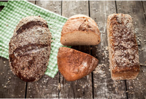 Auf einem Holzuntergrund liegen drei unterschiedliche rustikale Brote. Sie sind umgeben von Mehl und Körnern sowie von einem grün karierten Tuch. Das mittlere Brot ist angeschnitten und offenbart eine schöne gleichmäßige Krume.