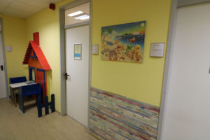 In der Logoädie-Praxis hängt ein buntes Bild mit Bärchen an einer gelben Wand. In einer Kinderecke steht ein Tisch mit blauen Stühlen. An der Wand ist eine bunte Hausfront in Kindergröße als Dekoration montiert.