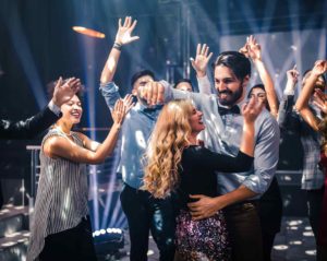 Eine Gruppe attraktiver junger Leute tanzt mit Freude. Ein Paar im Mittelpunkt zeigt Paartanz. Die Männer tragen Hemden, die Frauen Blusen oder Kleider. Einige klatschen oder jubeln. Eine Gruppe attraktiver junger Leute tanzt mit Freude. Ein Paar im Mittelpunkt zeigt Paartanz. Die Männer tragen Hemden, die Frauen Blusen oder Kleider. Einige klatschen oder jubeln.