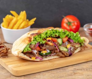 Auf einem Holzbrett liegt ein üppig gefülltes Fladenbrot mit Salat, Dönerfleisch, Tomate, Gurke, Mais und roten Zwiebeln. Im Hintergrund sind eine Tomate sowie Schälchen mit Pommes frites und Ketchup zu erkennen. Auf einem Holzbrett liegt ein üppig gefülltes Fladenbrot mit Salat, Dönerfleisch, Tomate, Gurke, Mais und roten Zwiebeln. Im Hintergrund sind eine Tomate sowie Schälchen mit Pommes frites und Ketchup zu erkennen.