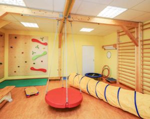 In einem Sportraum sind zahlreiche Geräte und Hilfsmittel für die Ergotherapie aufgebaut, darunter Kletterwände, eine Therapieschaukel, ein Kriechtunnel und ein Trampolin.