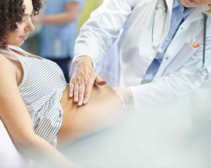 Eine Ärztin in weißem Kittel untersucht tastend den Schwangerschaftsbauch einer jungen Frau, die zurückgelehnt sitzt und lächelnd auf ihren Bauch blickt.