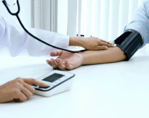 Eine Ärztin in weißem Kittel misst bei einer Person mithilfe eines Stethoskops den Blutdruck. Von beiden sind nur die Unterarme zu sehen.