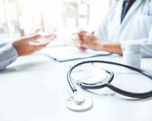Auf einem weißen Tisch liegt ein Stethoskop. Im Hintergrund spricht ein Mann mit einer Ärztin im weißen Kittel. Beide sitzen und sind nicht zu erkennen.