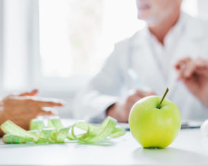 Auf einem weißen Tisch liegt ein grüner Apfel, daneben ein Maßband. Im Hintergrund führen zwei Personen sitzend ein Gespräch, sie sind nicht zu erkennen.