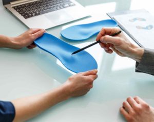 Über einem Tisch mit Glasplatte halten zwei Frauenhände eine blaue orthopädische Einlage. Eine zweite Person erläutert die Form der Einlage, indem sie mit einem Stift darauf zeigt.