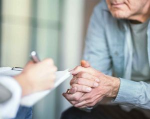 Eine Frau, von der man nur die rechte Hand sieht, macht Notizen auf einem Klemmbrett. Ihr gegenüber sitzt ein älterer Mann mit gefalteten Händen.