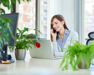 In einem Büro mit vielen Grünpflanzen sitzt eine junge Frau an einem Schreibtisch, schaut auf einen Laptop und telefoniert mit einem Smartphone.