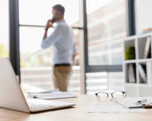 Auf einem Tisch in einem Büro steht ein aufgeklappter Laptop, daneben liegen eine Brille, ein Buch, Papiere und ein Stift. Im Hintergrund steht ein Mann im Hemd, der mit einem Smartphone telefoniert.