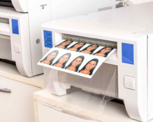 Aus einem weißen Drucker kommt ein Druckbogen mit biometrischen Passfotos einer dunkelhaarigen Frau.