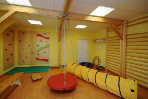 In einem Sportraum sind zahlreiche Geräte und Hilfsmittel für die Ergotherapie aufgebaut, darunter Kletterwände, eine Therapieschaukel, ein Kriechtunnel und ein Trampolin.