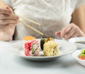 Eine Frau sitzt hinter einem Teller mit appetitlichen Sushirollen. Sie hält Essstäbchen in der rechten Hand. Neben dem Sushi stehen Schälchen mit Sojasauce, Wasabi und Sushiingwer. Eine Frau sitzt hinter einem Teller mit appetitlichen Sushirollen. Sie hält Essstäbchen in der rechten Hand. Neben dem Sushi stehen Schälchen mit Sojasauce, Wasabi und Sushiingwer.