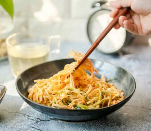 In einer dunkelgrauen Schale ist ein asiatisches Nudelgericht mit Gemüse angerichtet. Eine Person hebt einige Nudeln mit Essstäbchen an. Im Hintergrund steht eine Glastasse mit einem hellen Getränk. In einer dunkelgrauen Schale ist ein asiatisches Nudelgericht mit Gemüse angerichtet. Eine Person hebt einige Nudeln mit Essstäbchen an. Im Hintergrund steht eine Glastasse mit einem hellen Getränk.