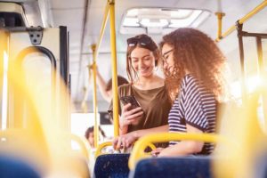In einem Linienbus mit gelben Stangen und Haltegriffen schauen zwei junge Frauen gemeinsam lächelnd auf ein Smartphone.