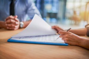 An einem Schreibtisch in einem Büro füllt eine Frau mit einem Stift ein Formular aus. Ihr gegenüber sitzt ein Mann im blauen Hemd, seine Hände sind gefaltet.