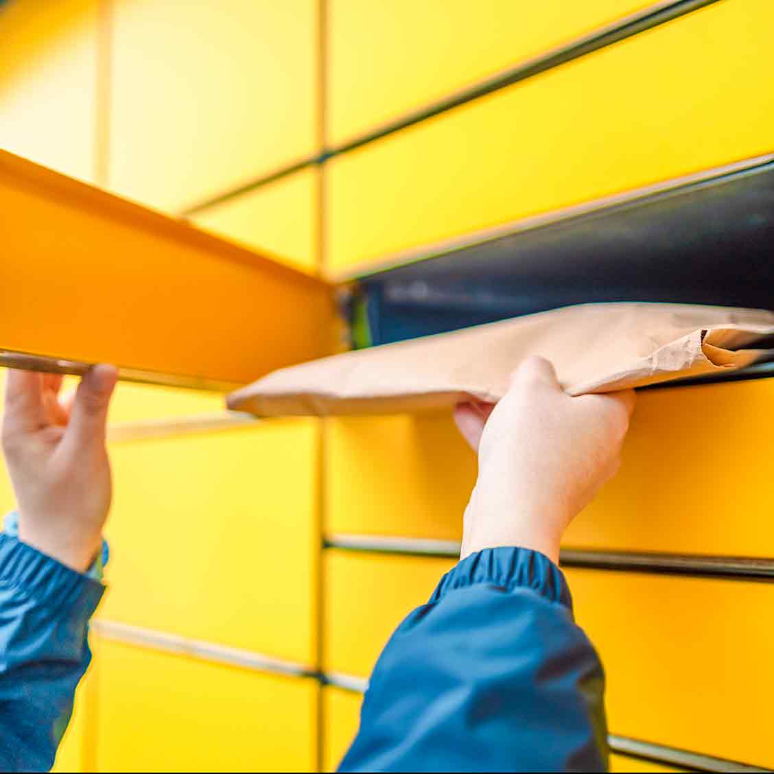 Eine Person, von der man nur die Unterarme sieht, steckt einen dicken braunen Umschlag in ein Paketfach einer gelben Poststation.