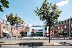 Über dem Haupteingang des Seidnitz Centers sehen wir blauen Himmel mit wenigen Wolken. Im Vordergrund befindet sich die Straße mit zwei Bäumen.