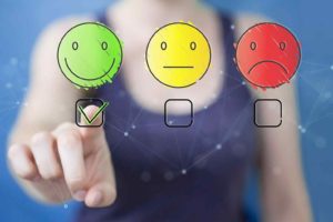 Auf einer durchsichtigen Fläche sind drei Smileys abgebildet: ein grünes Smiley mit lächelndem Mund, ein gelbes mit geradem Mund und ein rotes mit traurigem Mund. Hinter der Fläche steht eine unerkennbare Frau, welche mit dem Finger das grüne Smiley markiert.