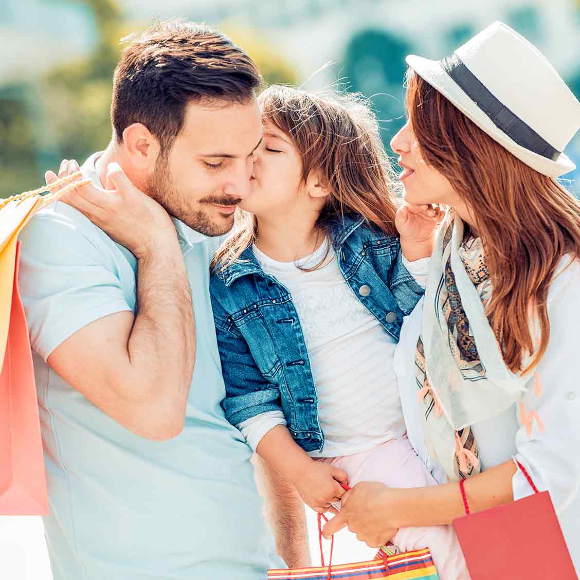Eine junge Familie in heller sommerlicher Kleidung hält Einkaufstaschen in den Händen. Die Mutter mit Hut hält die dreijährige Tochter auf ihrere Hüfte. Die Tochter gibt dem Papa einen Kuss auf die Wange.